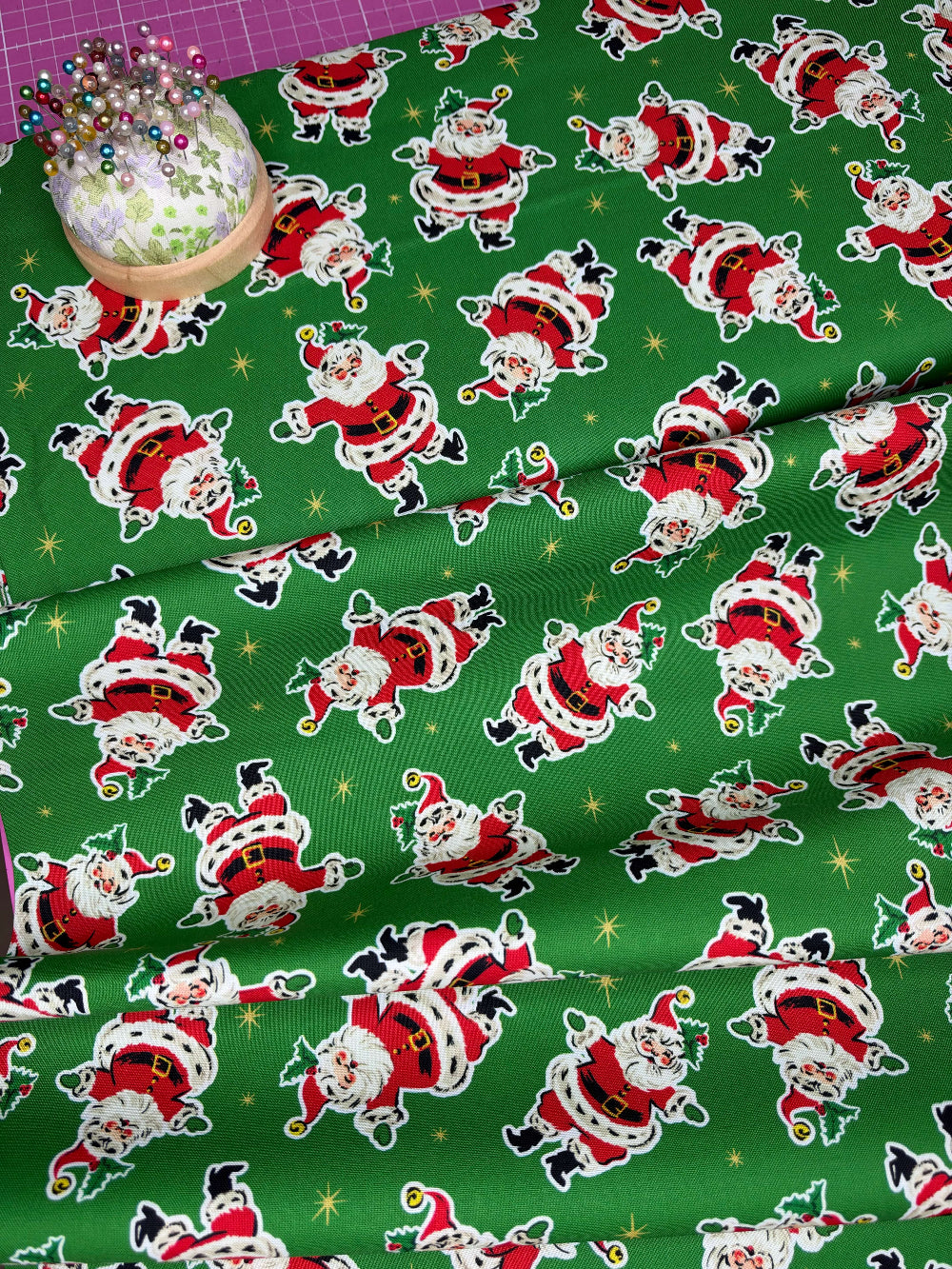 Green fabric with a repeating pattern of Santa Claus figures in red, white, and black, with small gold star accents. A pincushion with pins is visible in the top-left corner.
