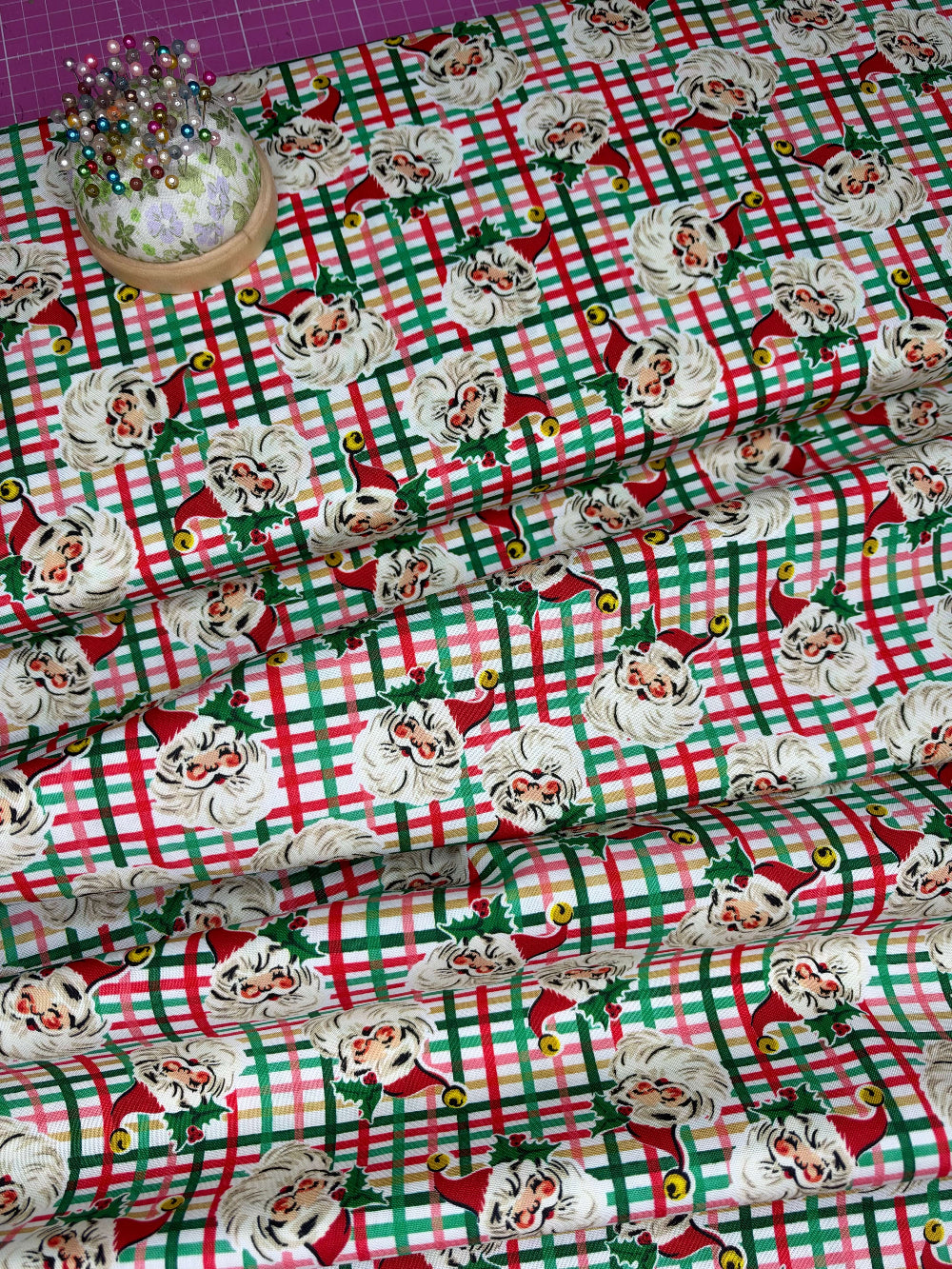 Christmas-themed fabric with a red, green, and white plaid pattern overlaid with Santa Claus faces, shown folded with a pin cushion in the corner.